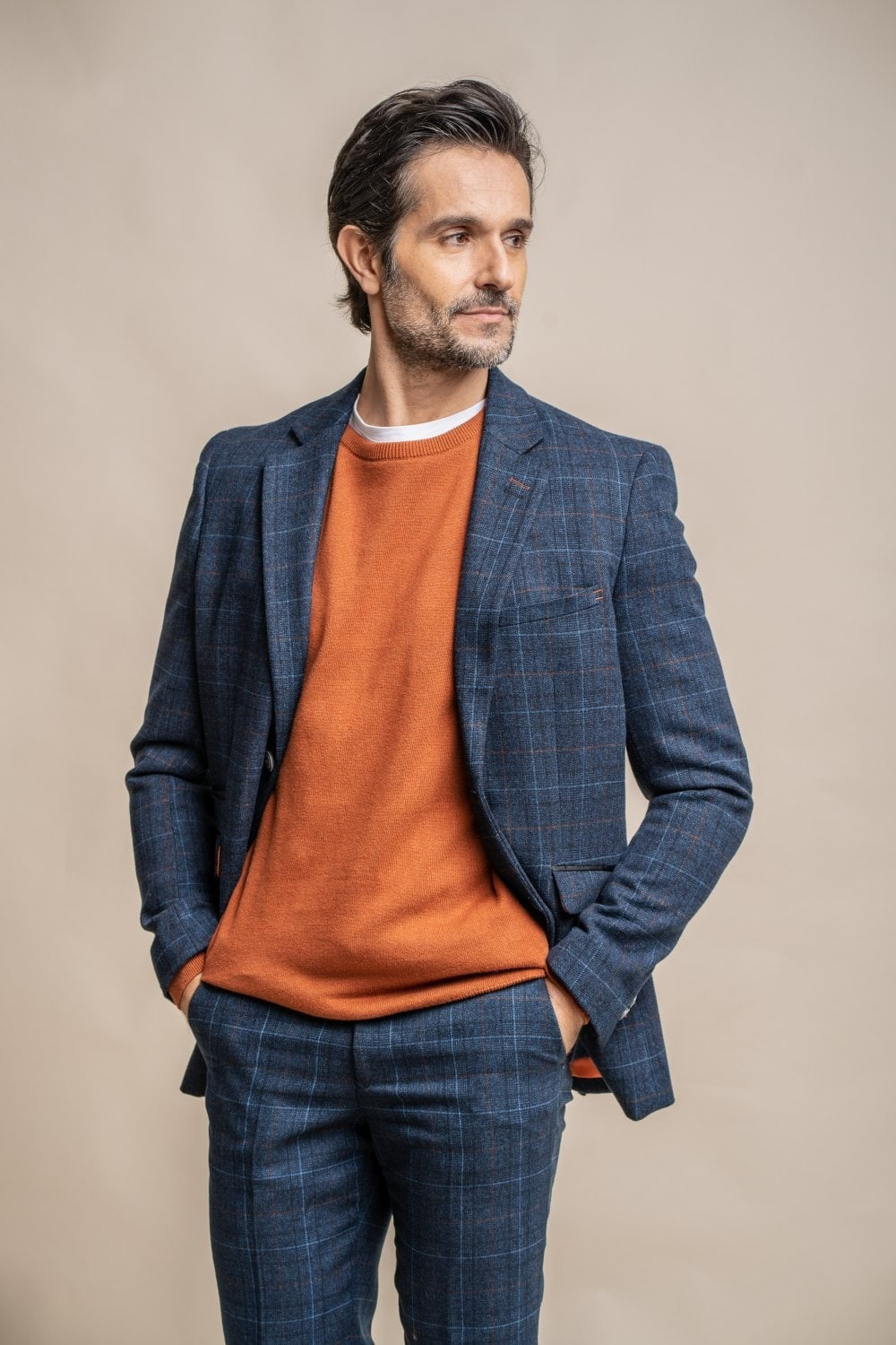 Man wearing a blue checkered blazer over an orange sweater and blue pants on a beige background